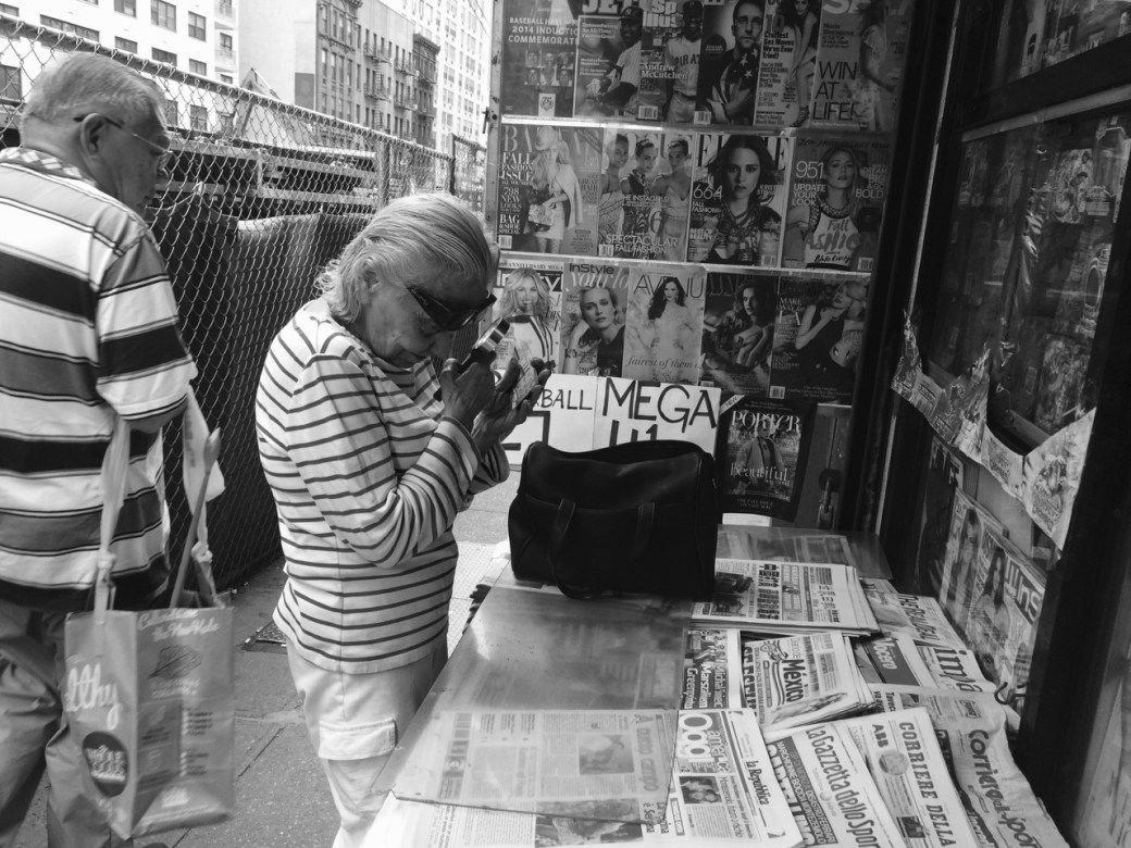 Woman at news stand peering at print through magnifying glass. Man walking by.