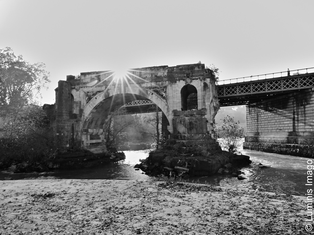 A monochrome image of the ruins an ancient roman bridge and the sun rising over them on a clear winter morning