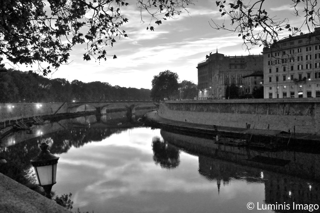 A black and white photograph of the river Tevere in Roma, with reflections of sky, trees and lights in its calm waters